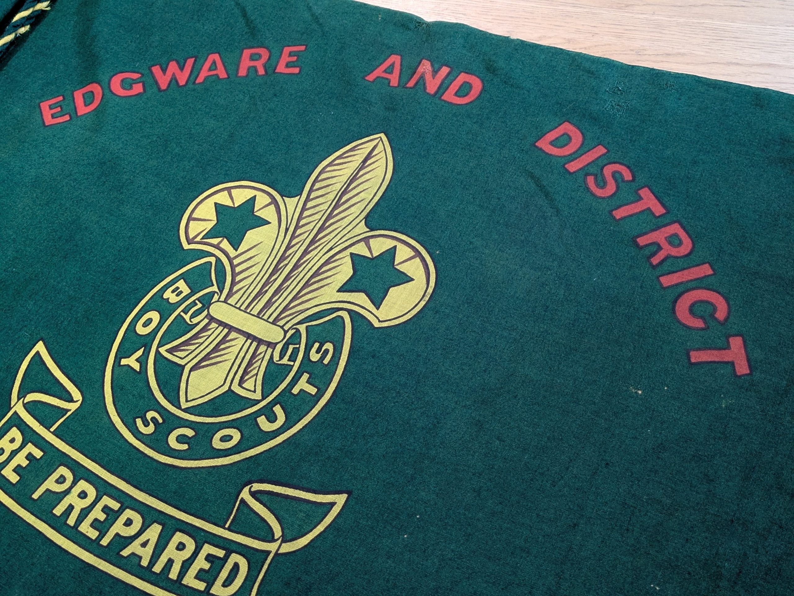 A green flag with 'Edgware and District' printed in red text, curved above a yellow Scout fleur-de-lis logo